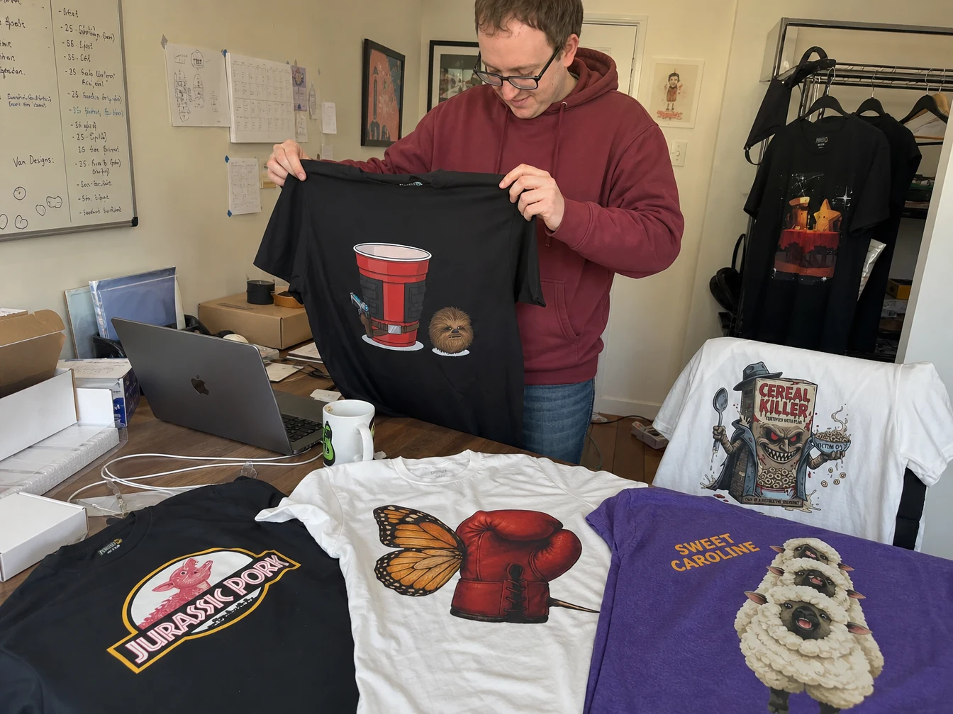 Jason holds a black Punish'd shirt while surrounded by several of the brand's T-shirt designs.